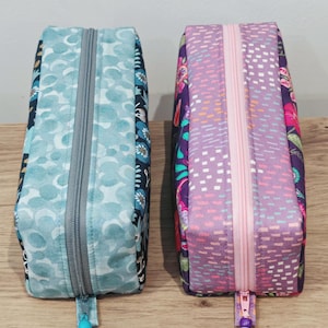 May include: Two rectangular fabric zip pouches. The left pouch features a light blue patterned top panel with a dark blue floral side. The right pouch has a light purple patterned top panel with a dark purple floral side. Both have beaded zip pulls.