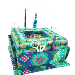 Può includere: Una scatola da cucito in tessuto verde acqua e verde con una varietà di strumenti da cucito, tra cui aghi, forbici e un porta spilli. La scatola ha un motivo a scacchi e un design floreale.