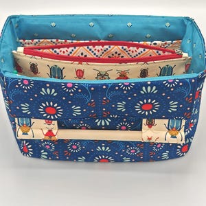 May include: A blue fabric organizer with a white handle and a floral pattern. The organizer has three compartments with zippers and a beige fabric lining. The compartments are decorated with a pattern of red and green beetles.