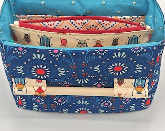 May include: A blue fabric organizer with a white handle and a floral pattern. The organizer has three compartments with zippers and a beige fabric lining. The compartments are decorated with a pattern of red and green beetles.