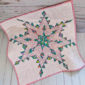 May include: A handmade quilt featuring a large starburst design. The star is composed of pink and floral patterned fabric, with a teal floral center. The quilt has a pink border and a light pink dotted background.