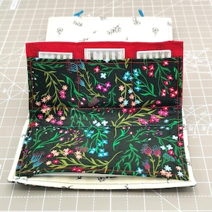 May include: A black fabric needle case with a floral pattern. The case has three compartments for storing needles. The case is open and the needles are visible.