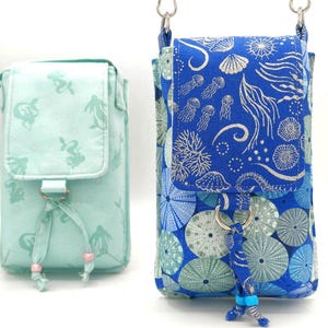 May include: Two fabric crossbody phone bags. One is light blue with a mermaid pattern, the other is blue with a sea life pattern. Both have a flap closure and a decorative tassel. The bags are designed to be worn over the shoulder.