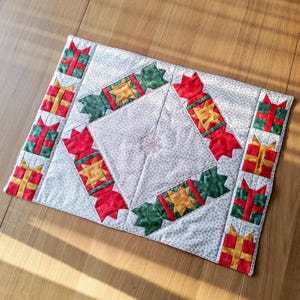 May include: A festive quilted placemat with a Christmas theme. It features a central diamond design with red, green, and gold cracker and gift box patterns. The background is light grey with small white polka dots and a snowflake design.