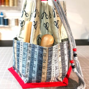 Puede incluir: Una bolsa de kit de costura hecha a mano con base y asa rojas. El exterior de la bolsa presenta tela estampada con tijeras y patrones de cinta métrica. Contiene herramientas de madera y un cordón gris con cuentas.