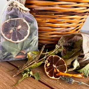 Peut inclure: Un sachet en voile lavande à cordon coulissant rempli de feuilles séchées et orné d'une tranche d'agrume séchée. À côté, un second sachet en toile de jute ouvert contient d'autres feuilles, une tranche d'agrume et des brins d'herbes séchées.