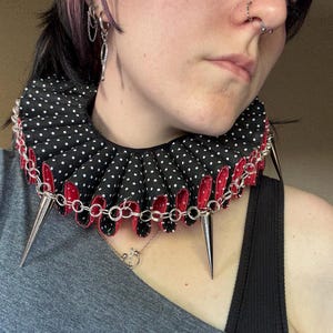May include: A black and white polka dot ruffled collar with red trim and silver chain accents. The collar features multiple silver spikes and is a unique fashion accessory.