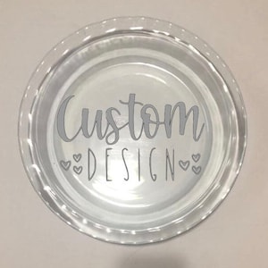 May include: A clear glass pie plate with the words "Custom DESIGN" in a gray script font. Small heart shapes are below the word "DESIGN". The pie plate has a fluted edge.