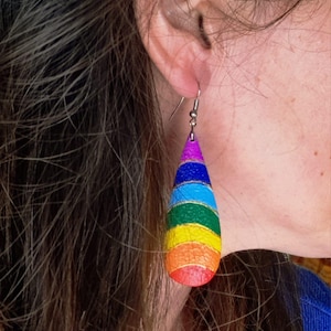 May include: Rainbow-striped teardrop earrings. The earrings feature horizontal bands of red, orange, yellow, green, blue, and purple. They are suspended from a silver hook. The earrings are made of a material that looks like leather or paper.