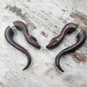 May include: A pair of dark brown wooden snake ear gauges. The gauges are carved in the shape of a snake's head and neck, with a curved tail. The gauges are made of wood and have a natural finish.