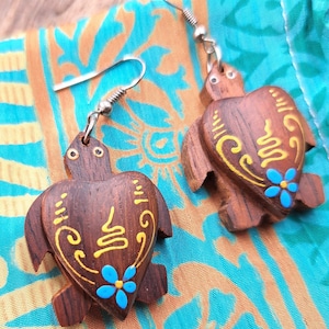 Peut inclure: Boucles d'oreilles en forme de tortue en bois, ornées de détails peints en jaune et bleu. Les boucles d'oreilles sont suspendues à des crochets d'oreilles argentés. Le fond est un tissu coloré avec des motifs orange et turquoise.