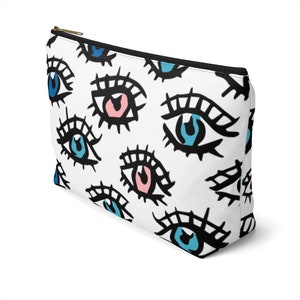 Könnte beinhalten: Weiße Kosmetiktasche mit Reißverschluss und einem sich wiederholenden Muster aus schwarz umrandeten Augen mit blauen, rosa und türkisfarbenen Iris.