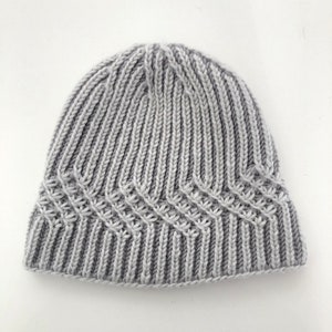 Puede incluir: Un gorro de punto gris con un patrón de canalé y un diseño texturizado. El gorro tiene un borde doblado.