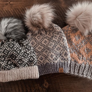 Puede incluir: Tres gorros de invierno de punto con pompones de piel sintética. Los gorros son de patrón blanco y negro, blanco y gris, y gris y melocotón.