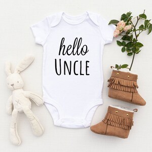 May include: White baby onesie with the text "hello uncle" in black script font. The onesie is lying on a white surface with a small stuffed bunny and brown baby booties.