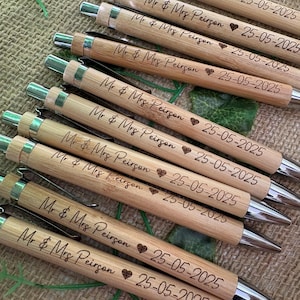 May include: Several bamboo pens, each inscribed with "Mr & Mrs Peirgon", a heart, and the date "25-05-2025". The pens feature silver metal components and are displayed on a textured background, suitable for wedding gifts.