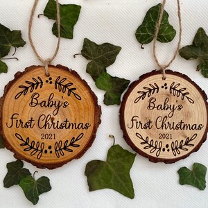 以下が含まれることがあります： 「Baby's First Christmas 2021」の文字と装飾的な葉のデザインが施された木製のオーナメント2個