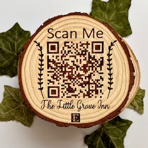 Puede incluir: Un disco redondo de madera con un patrón de veta de madera marrón. El disco tiene un código QR en blanco y negro con el texto "Scan Me" arriba y "The Little Grove Inn" abajo.