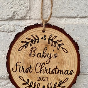 以下が含まれることがあります： 「Baby's First Christmas 2021」の文字と装飾的な葉のデザインが施された木製のクリスマスオーナメントです。