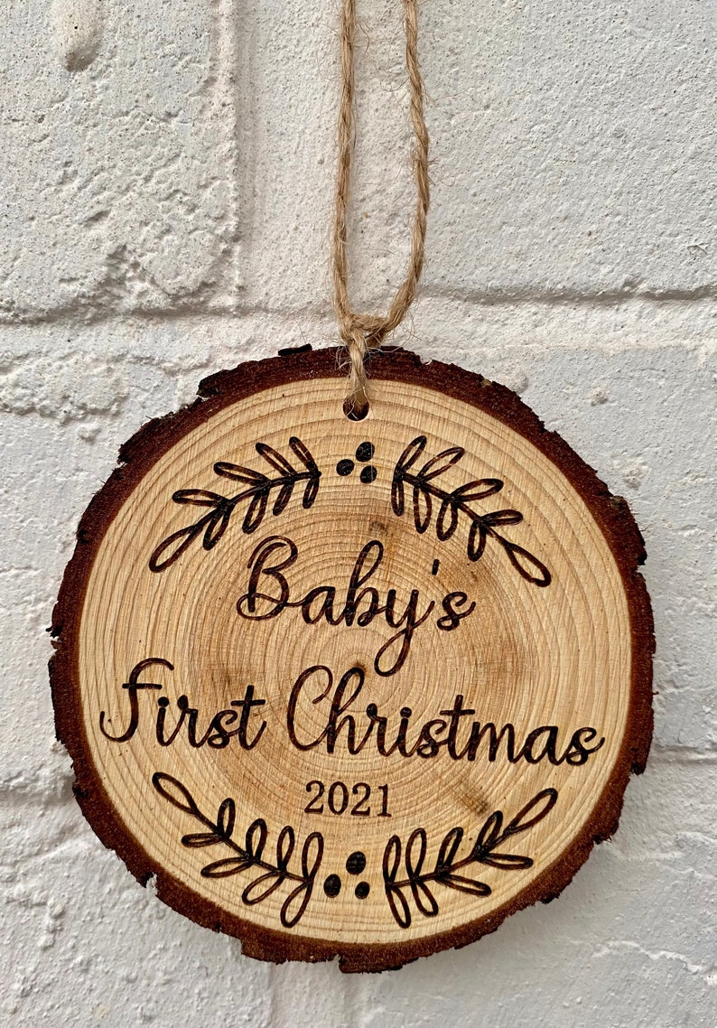 以下が含まれることがあります： 「Baby's First Christmas 2021」の文字と装飾的な葉のデザインが施された木製のクリスマスオーナメントです。
