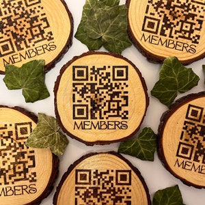 May include: Five wooden coasters with a laser-etched QR code and the word "MEMBERS" on each. The coasters are arranged in a circle on a white surface with green leaves scattered around them.