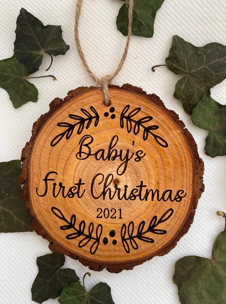 以下が含まれることがあります： 「Baby's First Christmas 2021」の文字と装飾的な花のデザインが施された木製のクリスマスオーナメントです。