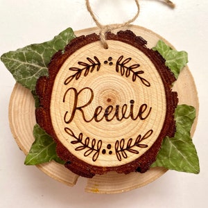 May include: A circular wooden ornament with the name "Reevie" inscribed in a cursive font, framed by a leaf design. The ornament features a natural bark edge, green leaves, and a twine loop for hanging.