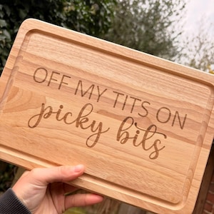May include: A rectangular wooden cutting board with a juice groove around the edge. The text "OFF MY TITS ON picky bits" is engraved on the board in a mix of block and cursive fonts. The board is held by a hand.