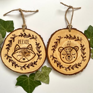 Peut inclure: Deux ornements en bois avec un motif de renard et d'ours. L'ornement de renard porte le nom "Reevle" et l'ornement d'ours porte le nom "JJ".
