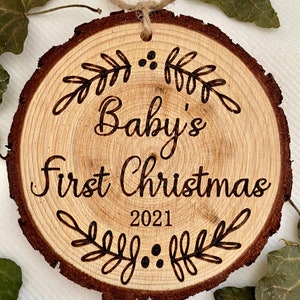 以下が含まれることがあります： 「Baby's First Christmas 2021」の文字と装飾的な葉のデザインが施された木製のクリスマスオーナメントです。