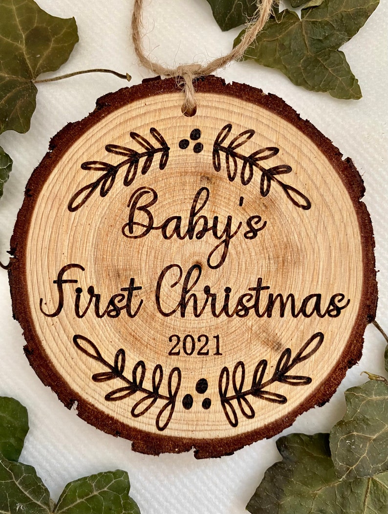 以下が含まれることがあります： 「Baby's First Christmas 2021」の文字と装飾的な葉のデザインが施された木製のクリスマスオーナメントです。