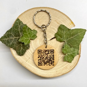 May include: A wooden keychain with a silver key ring and a laser-engraved QR code. The keychain is on a wooden surface with green leaves.