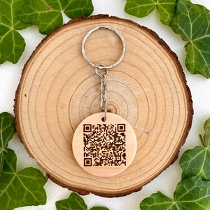 Peut inclure: Un porte-clés rond en bois clair avec un code QR gravé. Il est attaché à un anneau et une chaîne en métal argenté. Le fond est une tranche de bois entourée de feuilles de lierre vertes.
