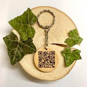 Op de afbeelding: Een houten sleutelhanger met een ronde, lichtbruine tag met een zwart-witte QR-code. De sleutelhanger is bevestigd aan een zilveren sleutelring en ketting.