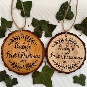 以下が含まれることがあります： 「Baby's First Christmas 2021」の文字と装飾的な葉のデザインが施された木製のオーナメント2つ。オーナメントは天然木で作られており、素朴な外観をしています。