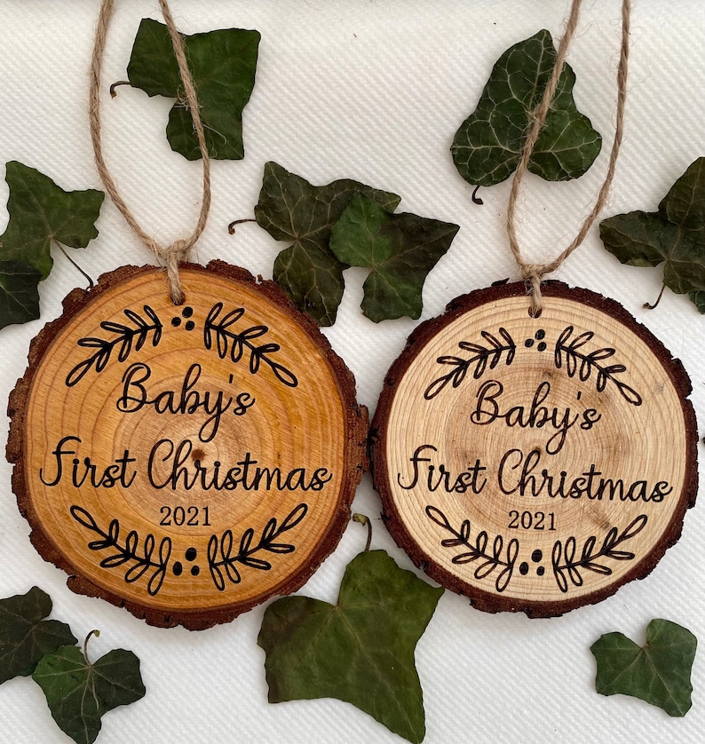 以下が含まれることがあります： 「Baby's First Christmas 2021」の文字と装飾的な葉のデザインが施された木製のオーナメント2つ。オーナメントは天然木で作られており、素朴な外観をしています。