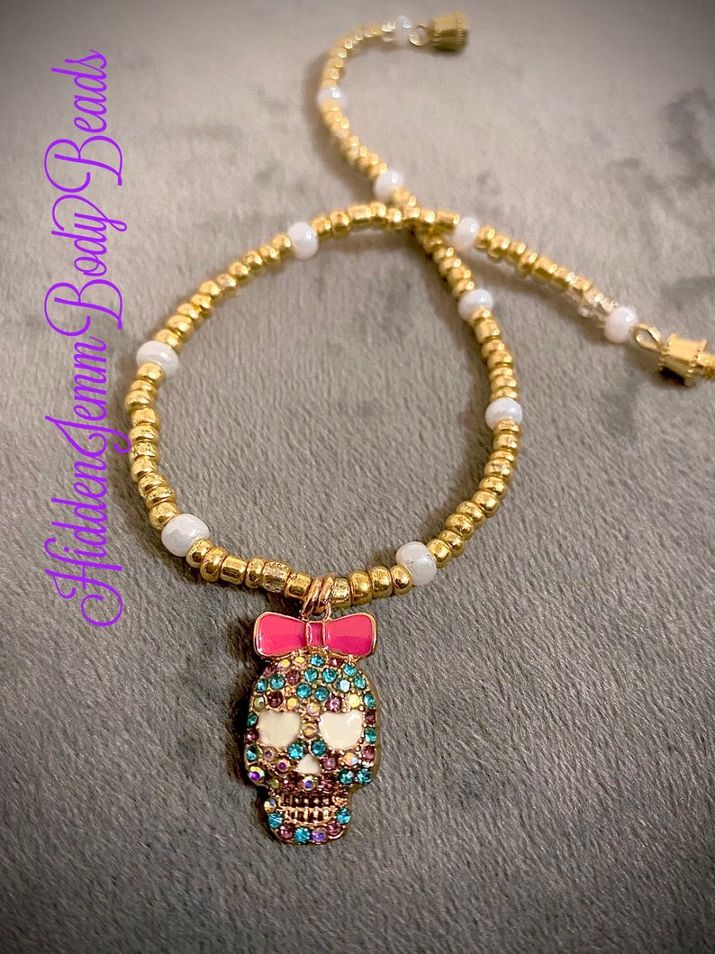 May include: A gold chain necklace with white beads and a pink bow charm featuring a jeweled skull. The skull is decorated with colourful rhinestones.
