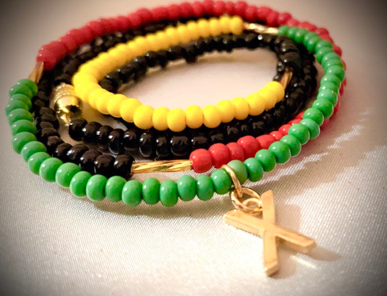 K&ouml;nnte beinhalten: Mehrschichtiges Perlenarmband mit roten, gelben, schwarzen und gr&uuml;nen Perlen. Das Armband hat einen goldenen Anh&auml;nger in Form eines Kreuzes.