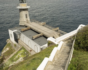 May include: A white lighthouse with a gray roof sits on a cliff overlooking the ocean. A set of stone steps lead up to the lighthouse, with a white railing along the side.