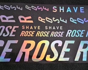 Könnte beinhalten: Ein rechteckiger schwarzer Aufkleberbogen mit mehreren Textzeilen in holografischer Ausführung. Der Text enthält das Wort ROSE, das in verschiedenen Größen und Ausrichtungen wiederholt wird, sowie das Wort SHAVE, das zweimal zu sehen ist.