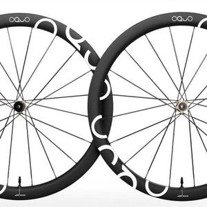 May include: A pair of black bicycle wheels with deep rims, featuring white OQUO branding printed along the outer edge. Each wheel has thin black spokes radiating from a central metallic hub, with a small valve stem visible at the bottom rim.