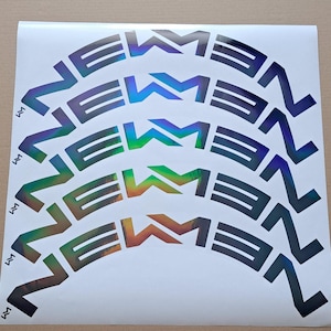 May include: A sheet of iridescent stickers with the word "NEWMEN" in a stylized font, arranged in a semi-circular pattern. The stickers have a metallic sheen, reflecting a spectrum of colors. Small "M" symbols are present.