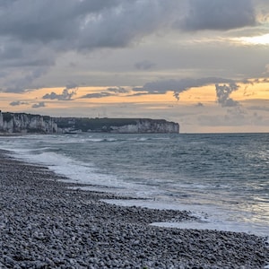 May include: A rocky beach with white cliffs in the distance. The sky is a mix of gray and orange clouds with the sun shining through.