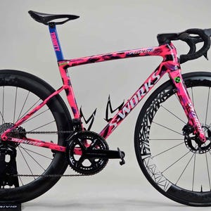 Könnte beinhalten: Ein rosa-schwarzes Specialized Rennrad mit schwarzen Rädern und einem schwarzen Sattel. Das Fahrrad hat ein rosa-schwarzes Tarnmuster auf dem Rahmen. Die Räder haben ein weißes geometrisches Design. Das Fahrrad steht auf einem schwarzen Ständer.