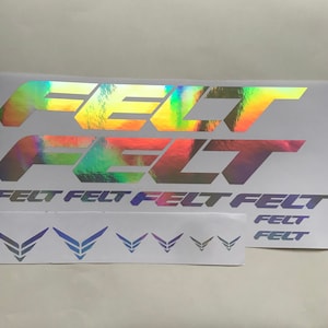 以下が含まれることがあります： 大胆で様式化されたフォントで「FELT」という言葉が書かれたホログラフィックステッカーのシート。ステッカーは虹色の光沢があり、さまざまな色を反射します。翼のようなデザインの小さなステッカーも含まれています。
