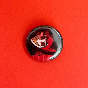 May include: A round button with a cartoon image of a red demon with black hair and a mischievous grin. The demon is wearing a red and black outfit with a heart-shaped design.