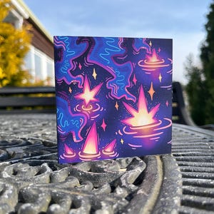 May include: Square art print featuring a vibrant, abstract design. The artwork showcases a cosmic theme with a dark blue background, accented by pink, purple, and yellow starbursts and swirling patterns. The print is displayed outdoors on a textured surface.