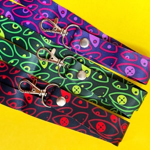 Könnte beinhalten: Vier bunte Lanyards mit einem sich wiederholenden Augenmuster. Jedes Lanyard hat einen silbernen Drehverschluss und einen silbernen Knopf.