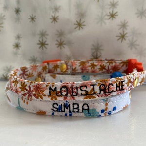 May include: Two fabric cat collars with adjustable buckles. The top collar is orange and white with the name "Moustache" embroidered in black. The bottom collar is white with a floral pattern and the name "Simba" embroidered in black.