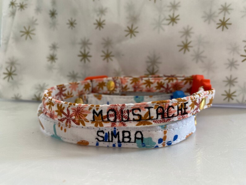 May include: Two fabric cat collars with adjustable buckles. The top collar is orange and white with the name "Moustache" embroidered in black. The bottom collar is white with a floral pattern and the name "Simba" embroidered in black.